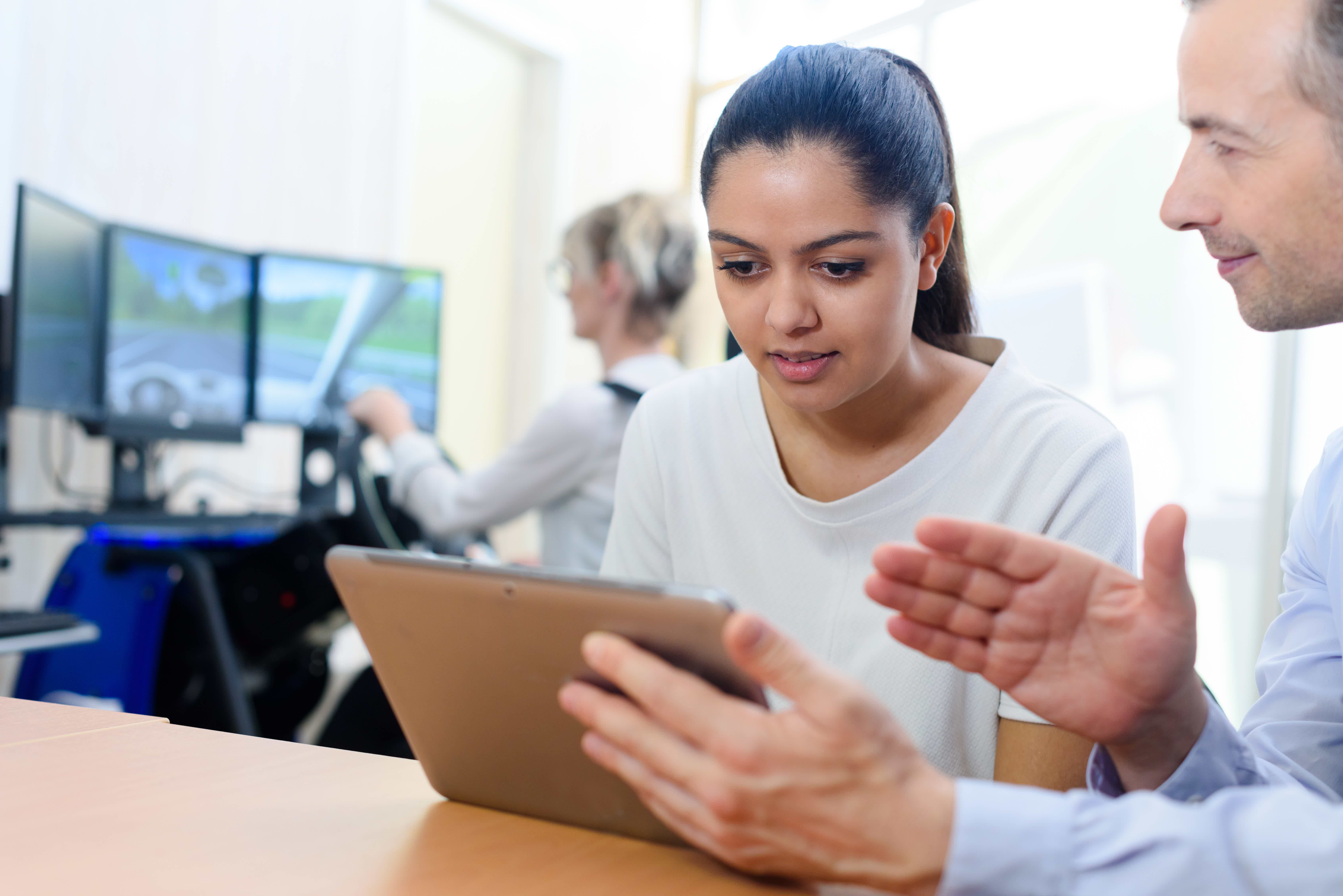 Mulher olhando tablet com pessoa fazendo aula em simulador de direção ao fundo