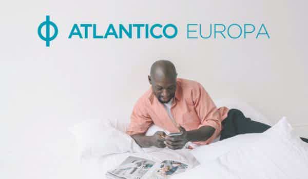À frente, logo do banco Atlantico Europa na parte superior da tela. Abaixo do logo vemos um homem deitado em uma cama com um telemóvel em sua mão e uma revista sobre o móvel.