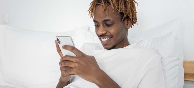 Homem sentado em uma cama observa a tela de seu celular.