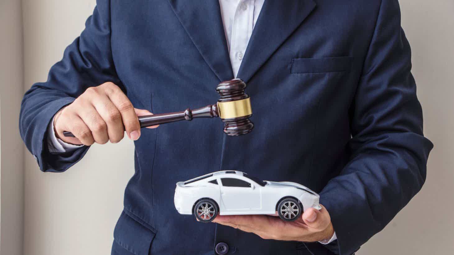 homem de terno azul bate um martelo de juiz sobre uma miniatura de carro branca em sua mão