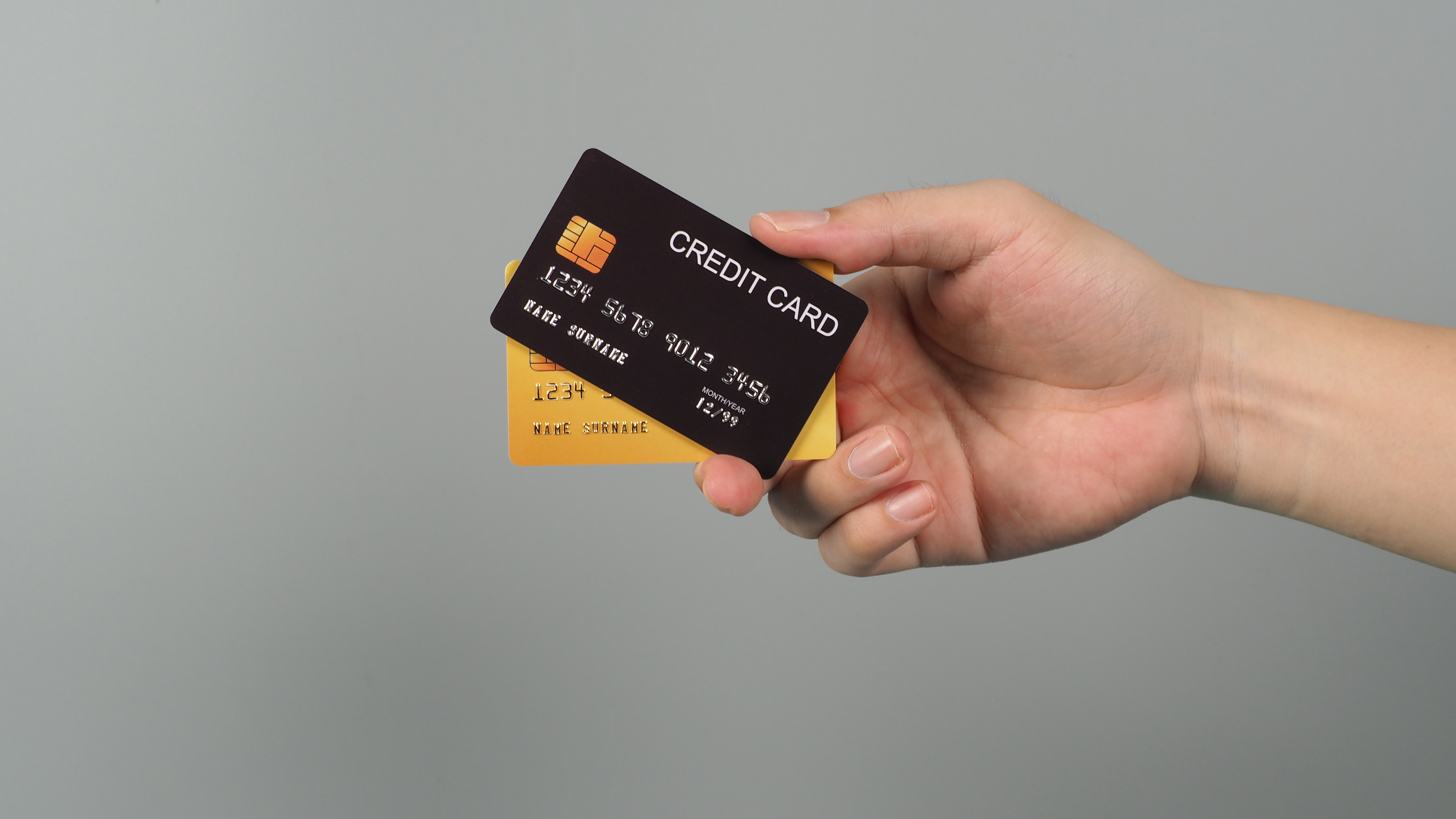 Na imagem mostra a mão de uma pessoa segurando um cartão preto e outro dourado em um fundo cinza