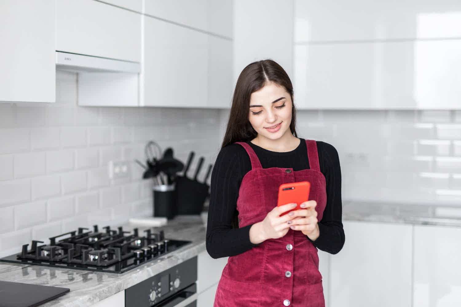 Menina com macacão bordô usando o celular na cozinha
