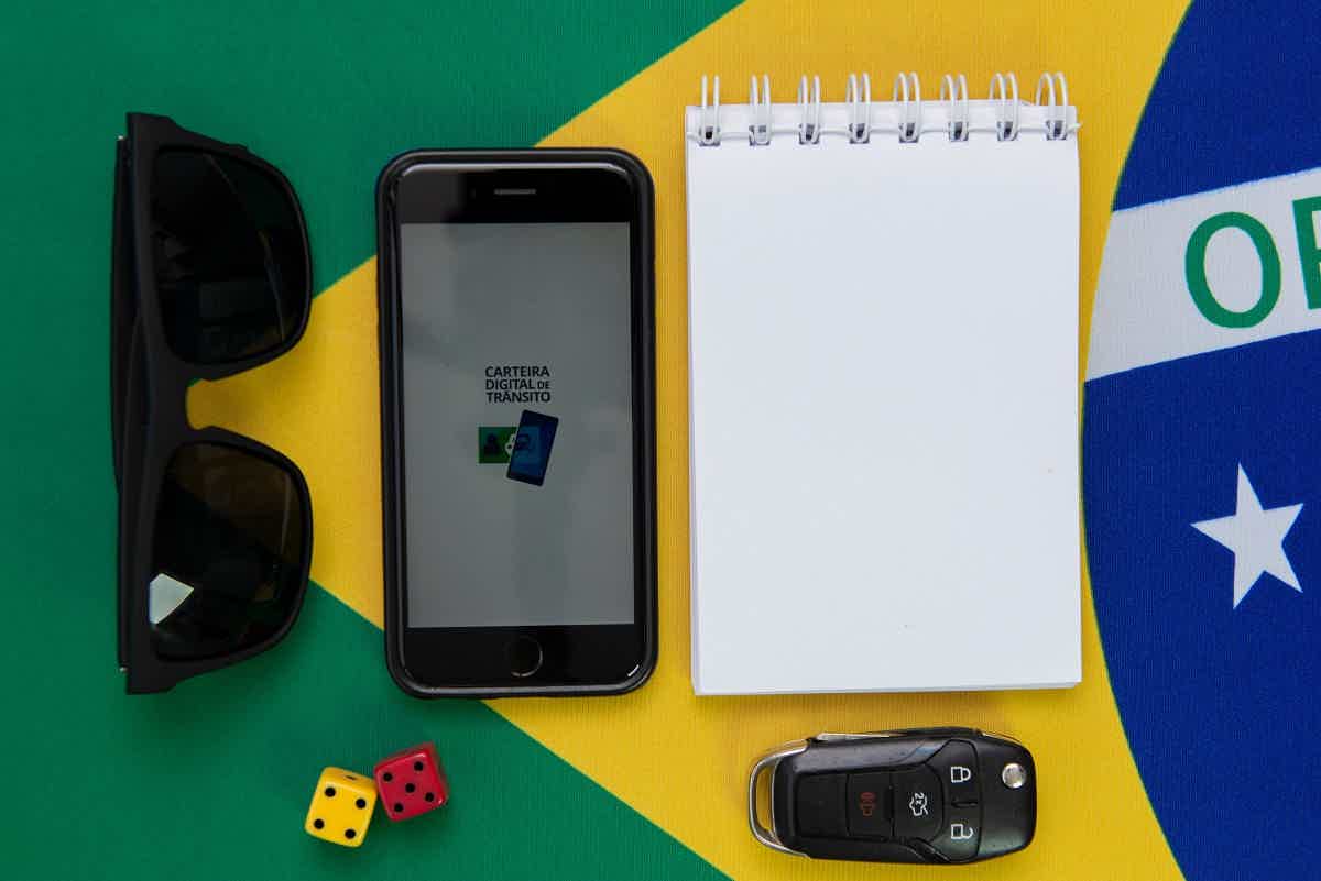 mesa com bandeira do brasil ao fundo; em cima dela: dados, óculos de sol, chave, bloco de anotações e celular aberto com aplicativo da CNH Digital