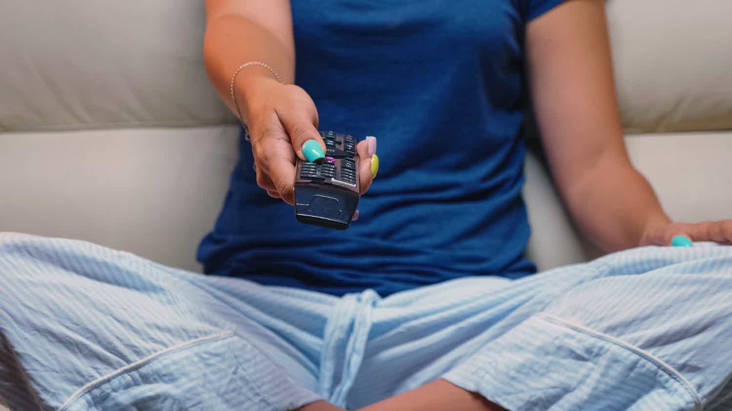Mulher usando pijama azul sentada em um sofá bege enquanto aponta o controle remoto para a TV