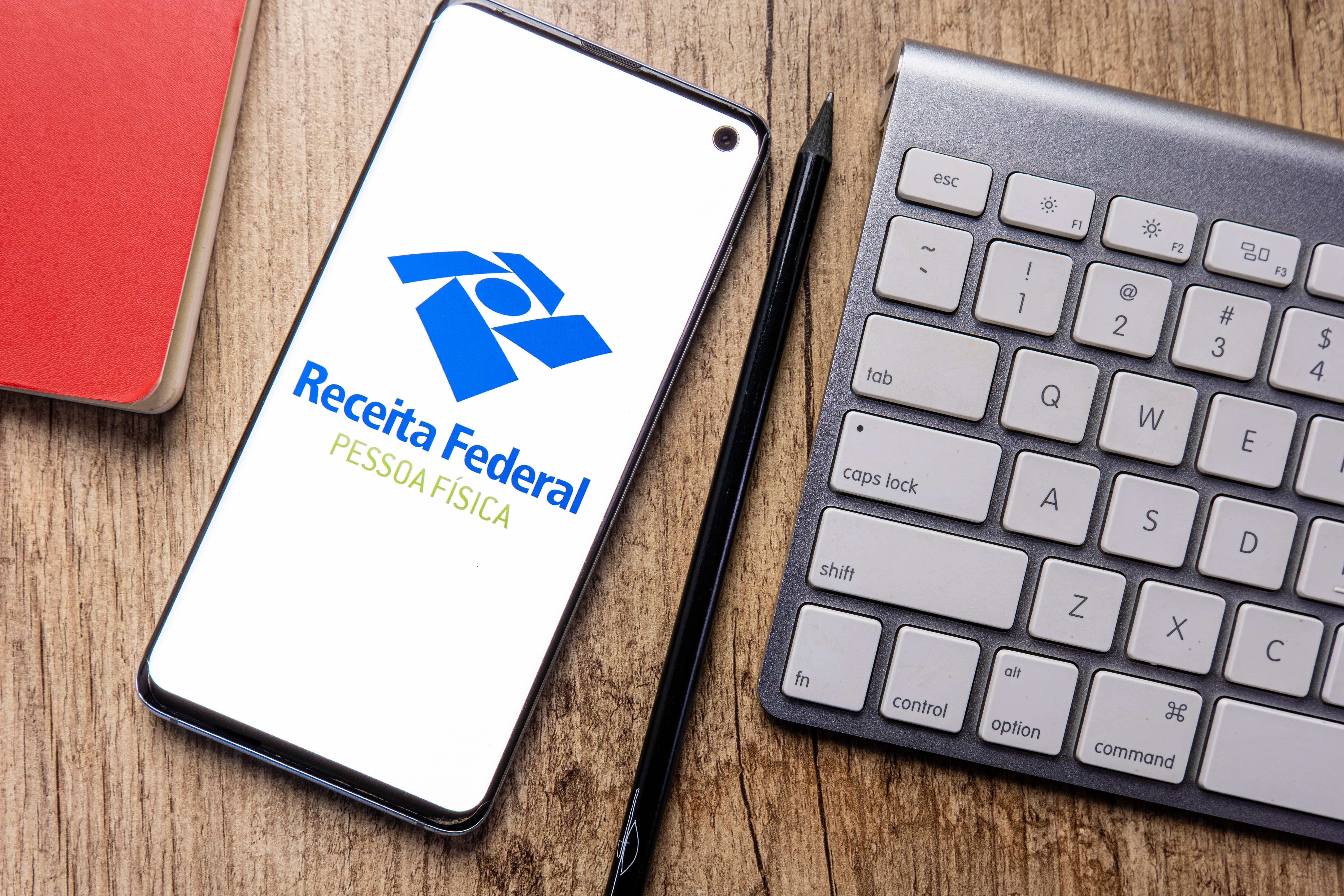 Imagem de um celular ao lado de um teclado repousado em uma superficie de madeira. O celular mostra a logo da receita federal.