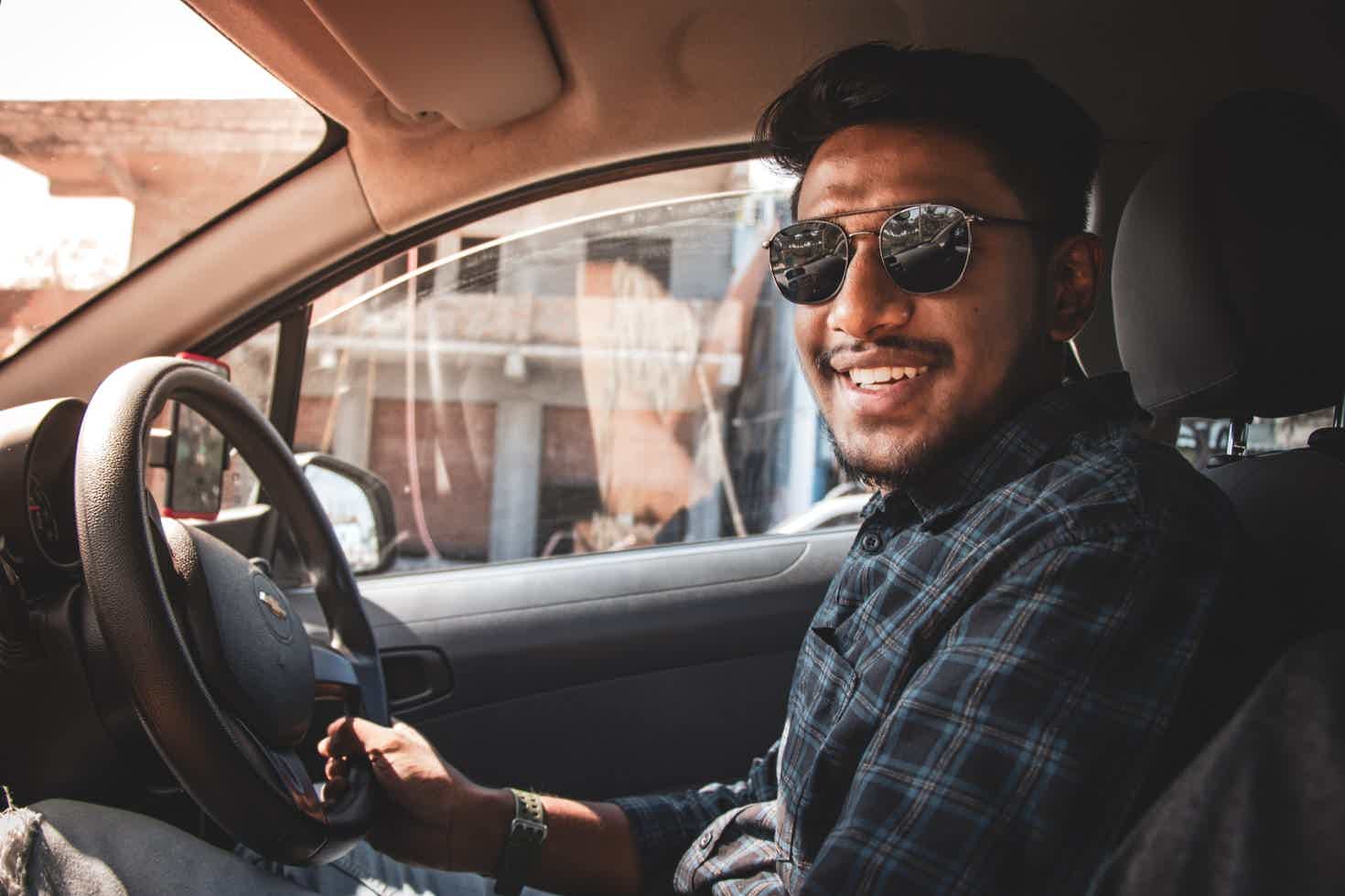 Pessoa de óculos escuros sentada no banco do motorista; sorrindo olhando para a câmera