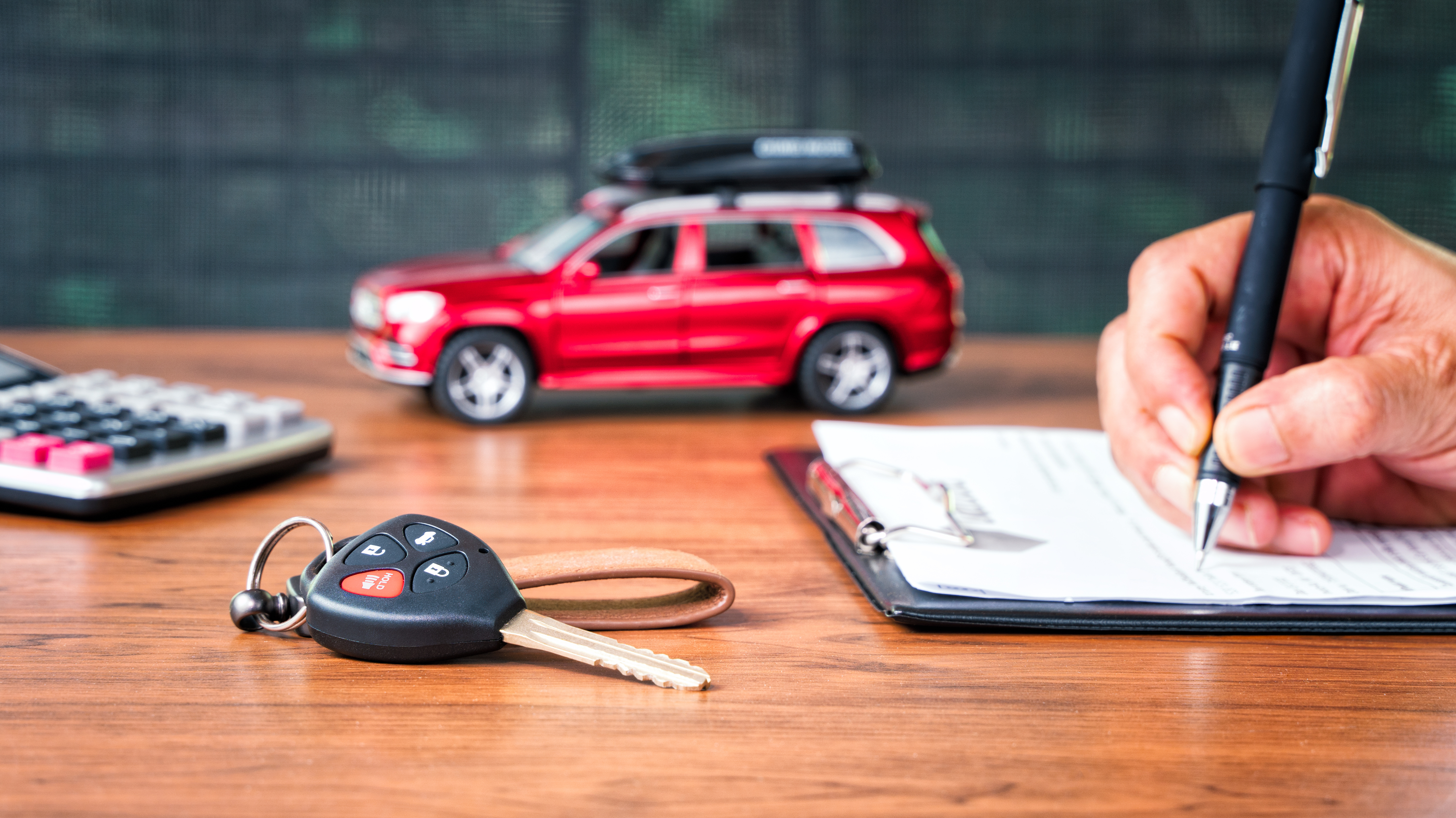 Mão assinando documento sobre mesa com chave de carro, calculadora e carro miniatura ao lado