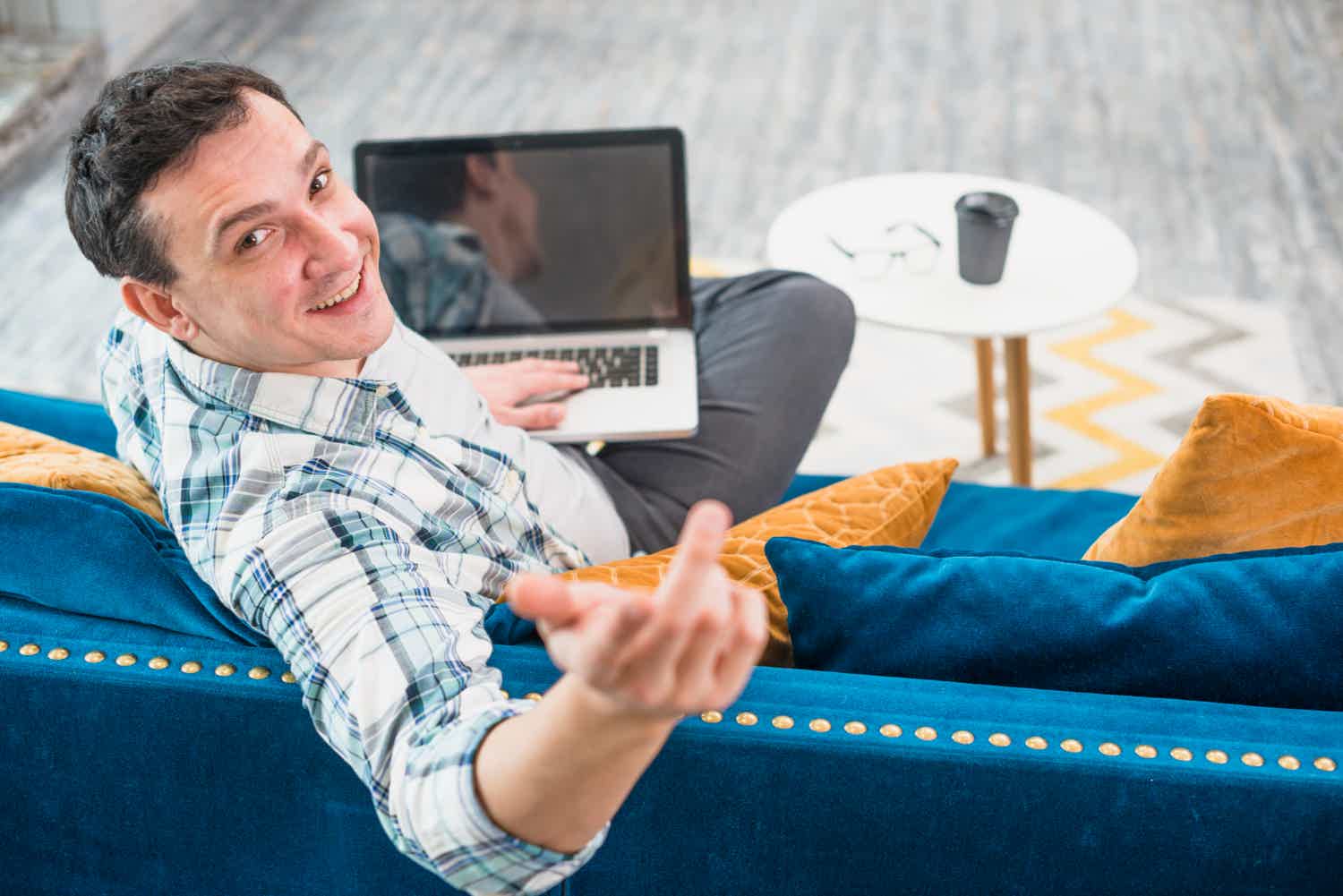 Homem sentado em sofá azul sorrindo com um notebook no seu colo