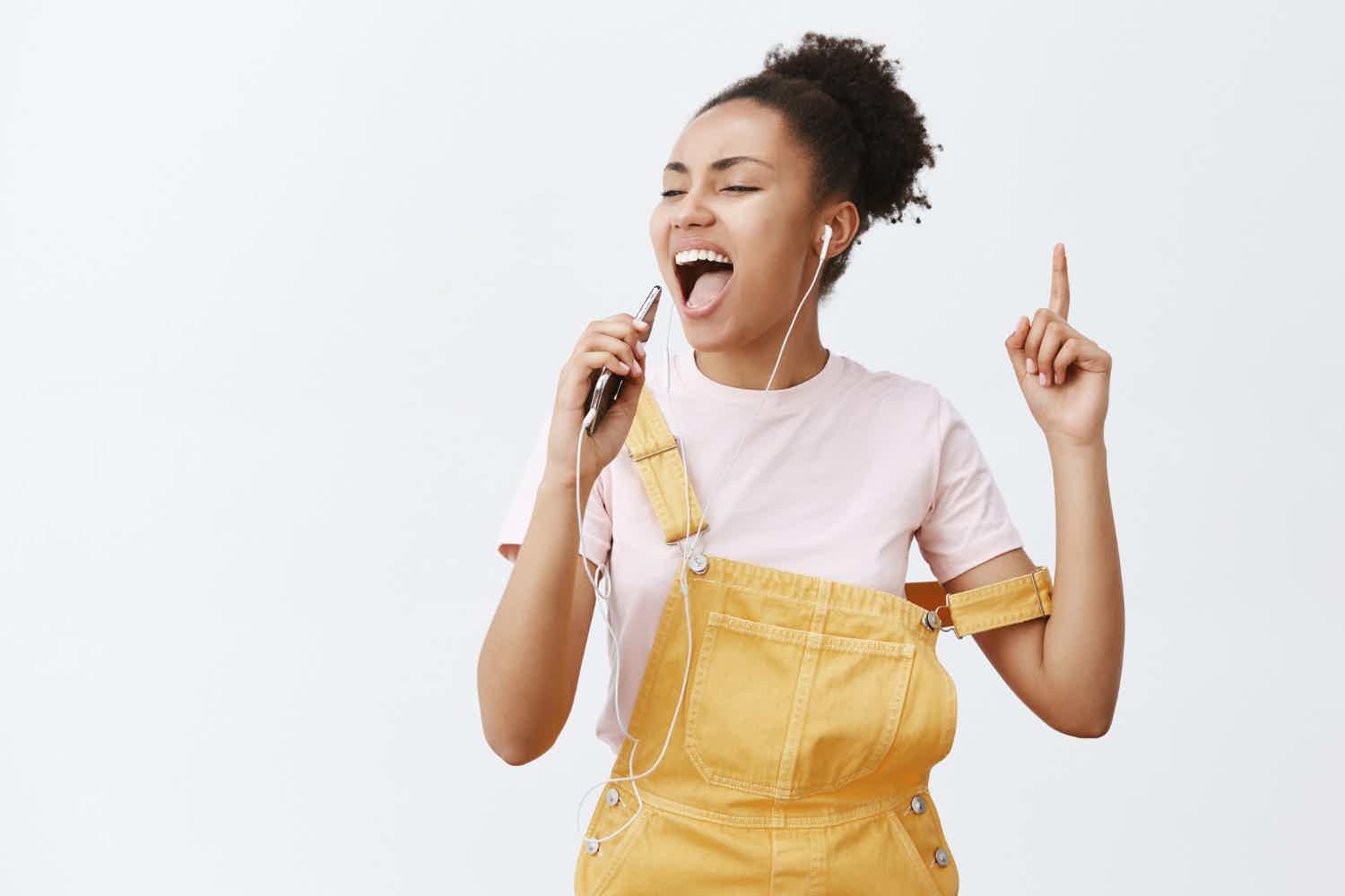 Menina negra usando macacão jeans amarelo e usando o celular de microfone enquanto canta