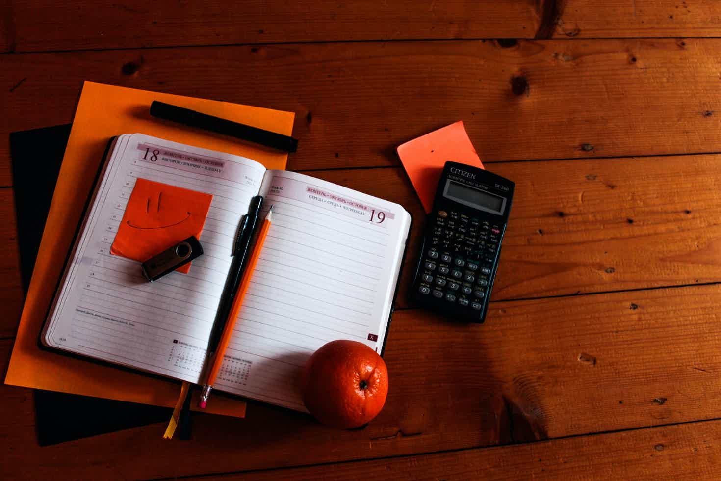 Caderno aberto, caneta, pen-drive e maçã em cima da mesa