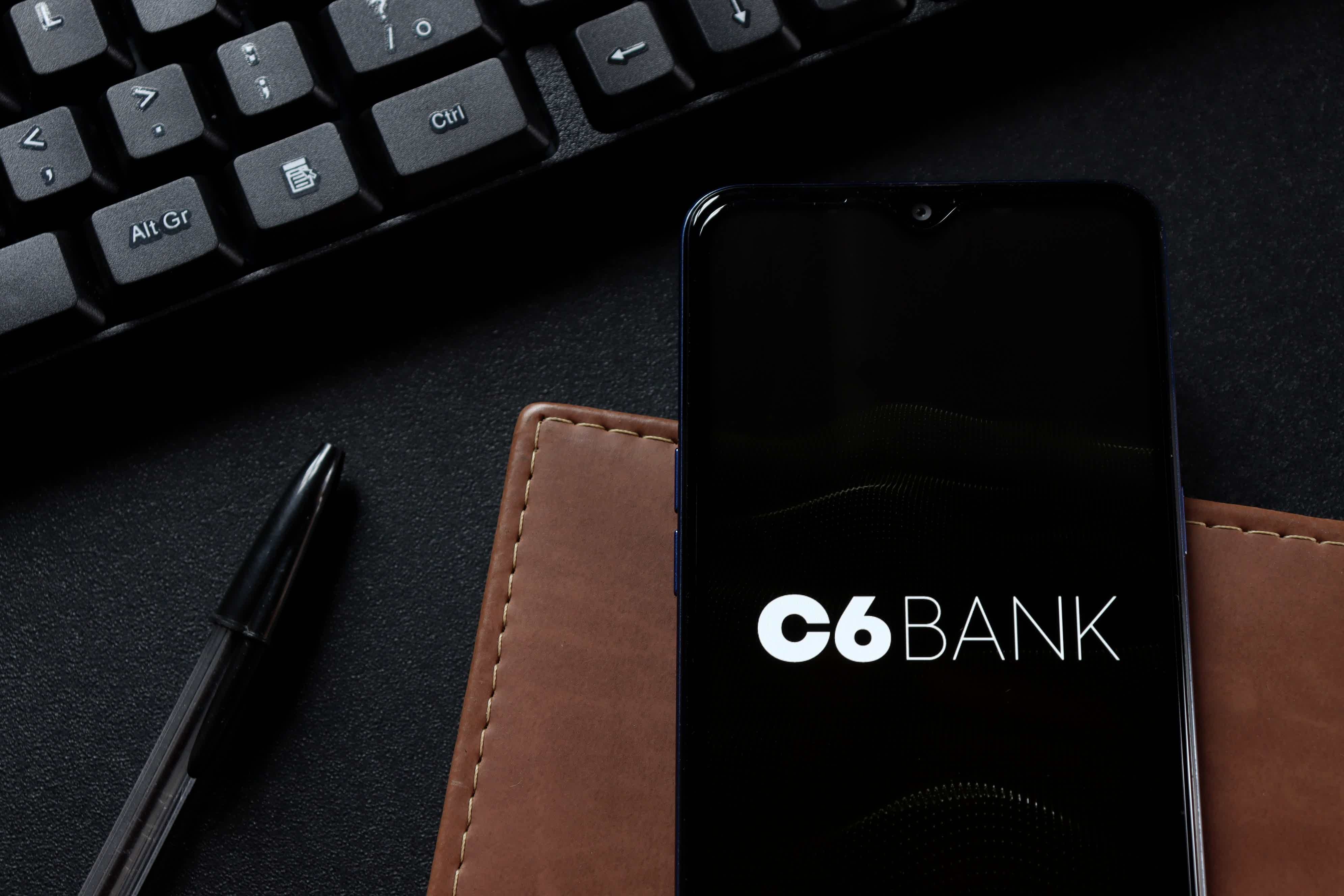 Imagem de celular com a logo do C6 Bank próximo a um teclado de computador e em cima de uma carteira marrom